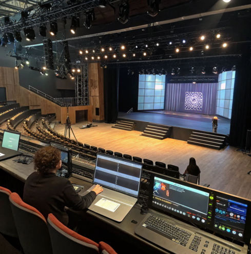 Un technicien contrôle l’éclairage et les écrans d’un grand théâtre vide depuis une régie, en face d’une scène illuminée.
